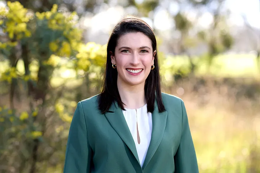Sian Keys, Chair of the Aspen Medical Foundation, wearing a green blazer and white blouse, smiling in a sunlit outdoor setting with soft-focus trees, yellow wattle flowers, and natural greenery in the background.
