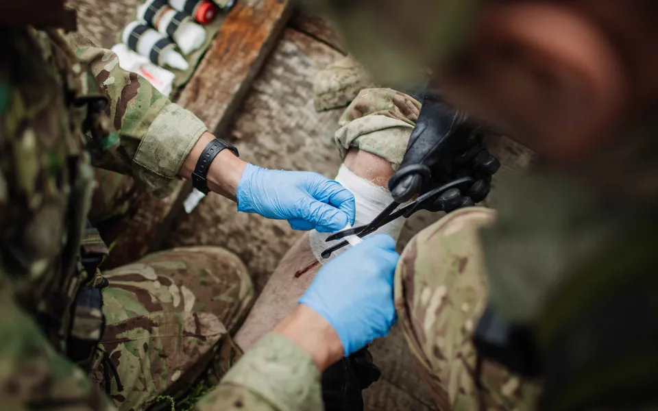 Image shows gloved hands cutting a bandage with people in defence uniforms.